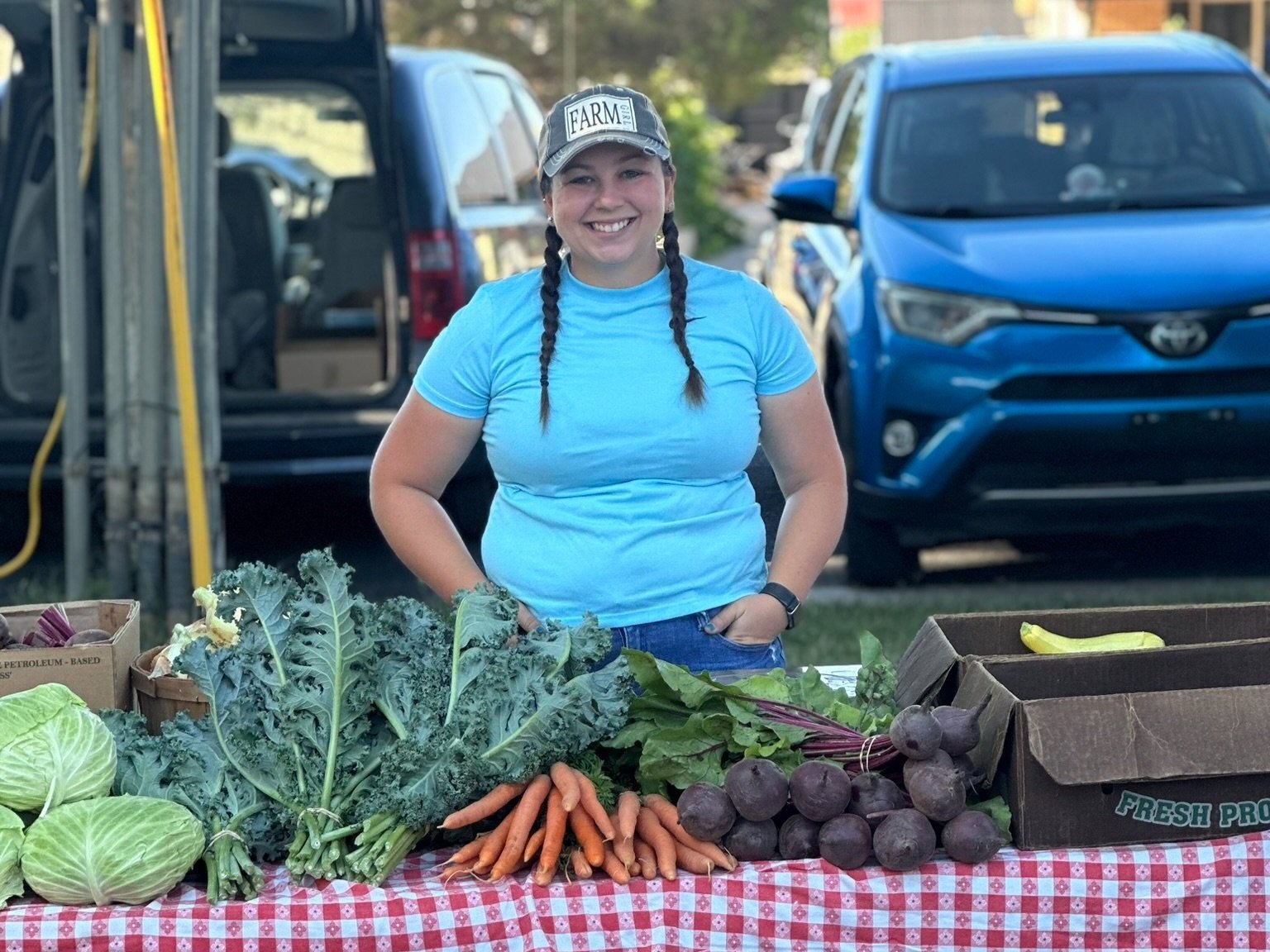 Smiling woman at a farmers market stand showcasing fresh vegetables including kale, carrots, beets, and cabbage, with a blue car in the background.